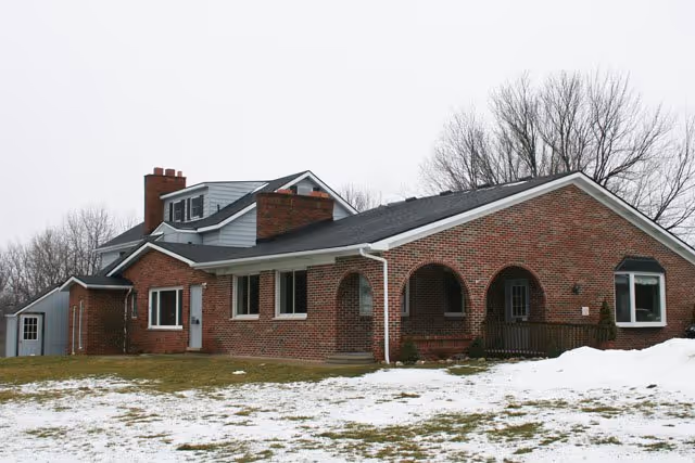Brick single-story house with arched front porch and a snow-dotted lawn under an overcast sky.