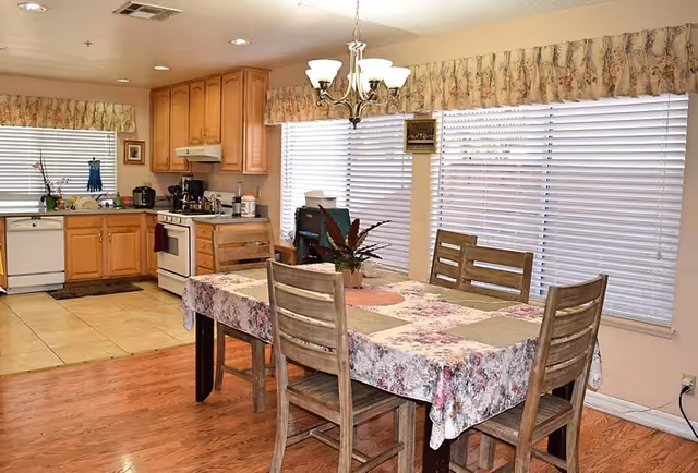 A residential kitchen and dining area with wooden cabinets, a stove, dishwasher, and countertop appliances. The dining table is covered with a floral tablecloth and surrounded by four wooden chairs. Large windows with blinds and floral valances let in natural light, and a chandelier hangs above the table.