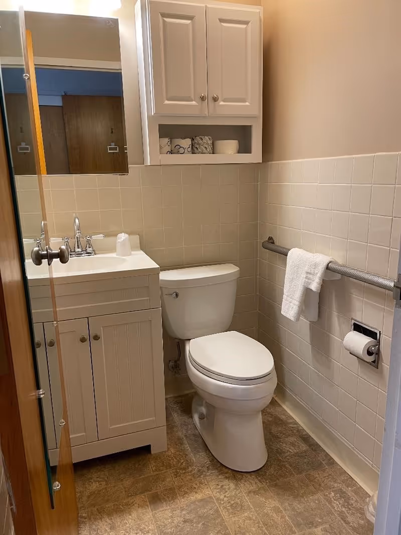 Compact bathroom featuring a toilet next to a sink vanity, a wall cabinet above, tiled half-walls, and a grab bar with a towel.
