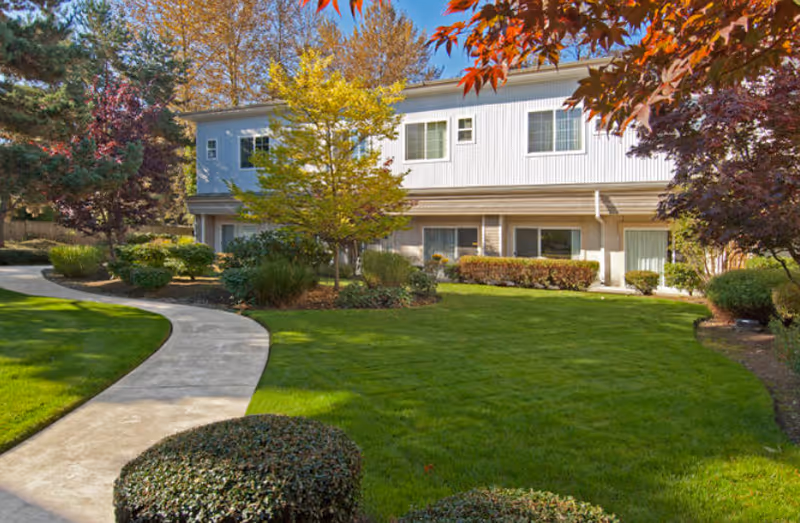 A curved sidewalk leads across a manicured lawn to the front of a two-story residential building surrounded by trees and shrubs.