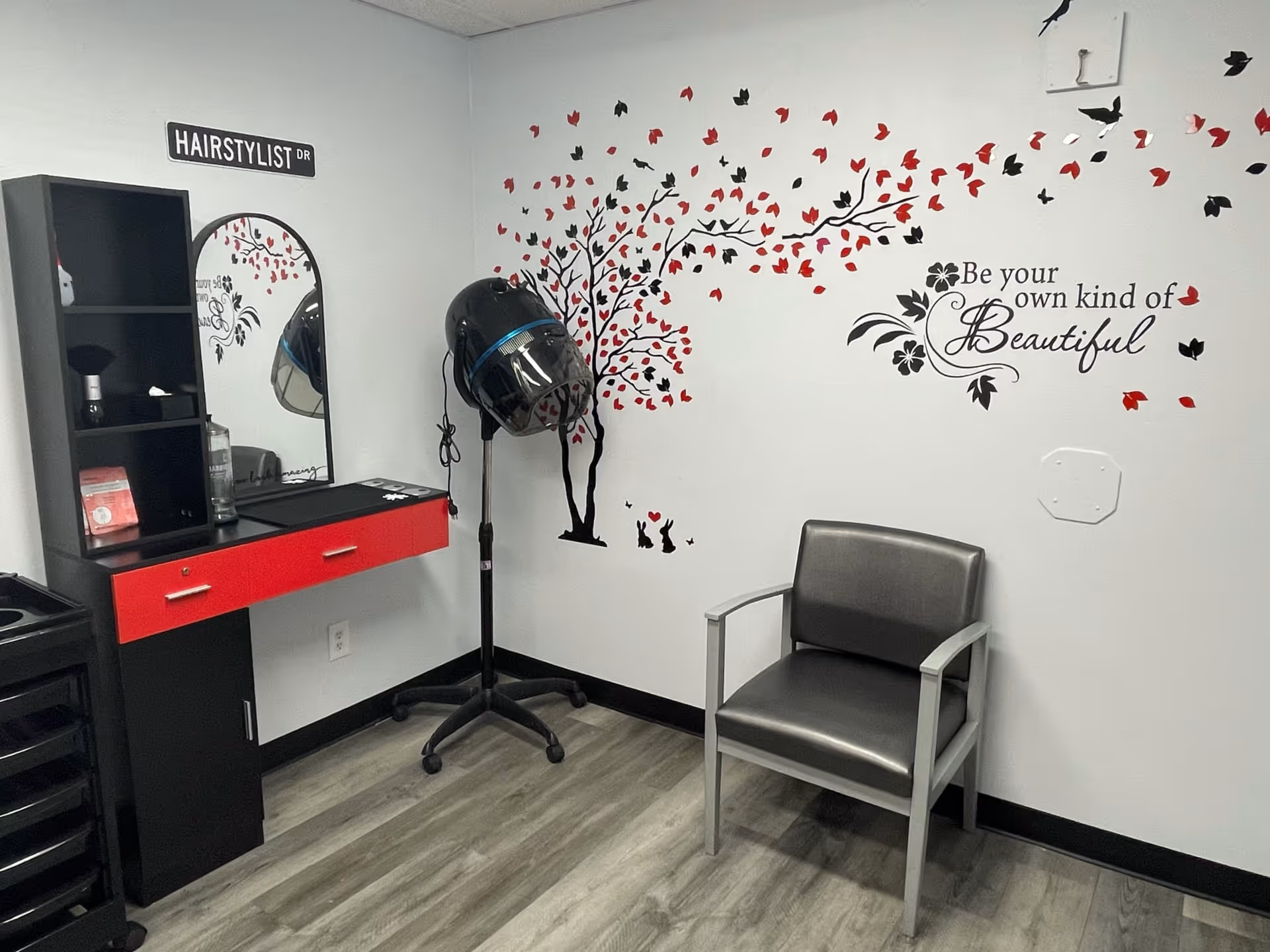 Interior of a hairstyling room with a black and red vanity table, a mirror, a black hair dryer on a stand, a gray chair, and a wall decal of a tree with red and black leaves along with the quote 'Be your own kind of Beautiful'.
