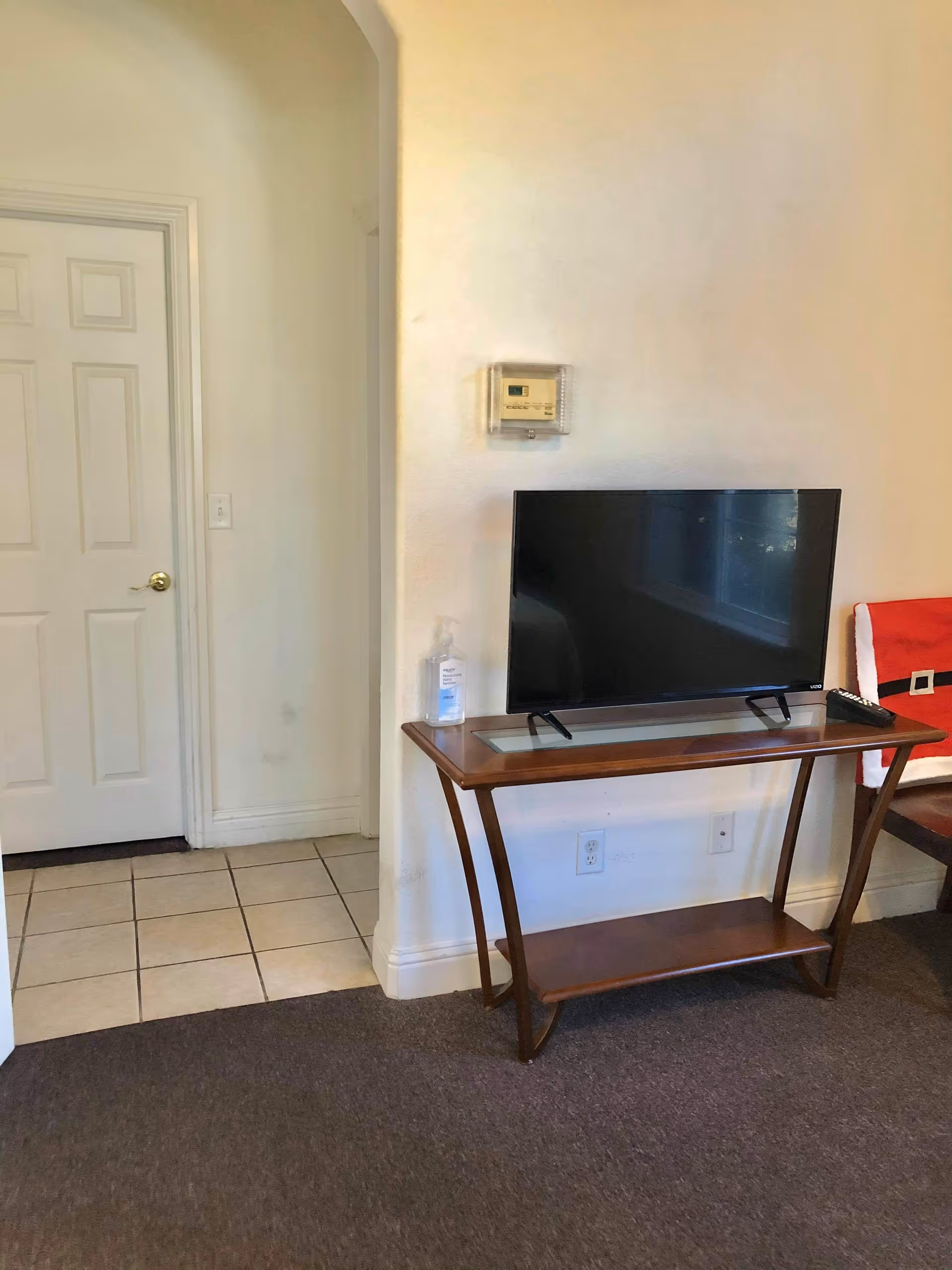 A small living area with a flat-screen TV on a wooden table. There is a bottle of hand sanitizer on the table next to the TV. A thermostat is mounted on the wall above the table. To the left, there is a doorway leading to a tiled floor area with a closed white door.