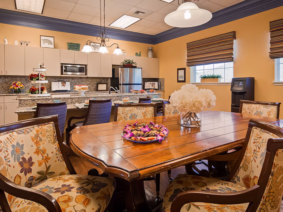 A warm and inviting kitchen and dining area with a wooden dining table surrounded by floral upholstered chairs. The kitchen features granite countertops, white cabinets, a stainless steel refrigerator, and a microwave. Decorative flowers and desserts are placed on the table and counter. Two windows with striped blinds allow natural light into the room.
