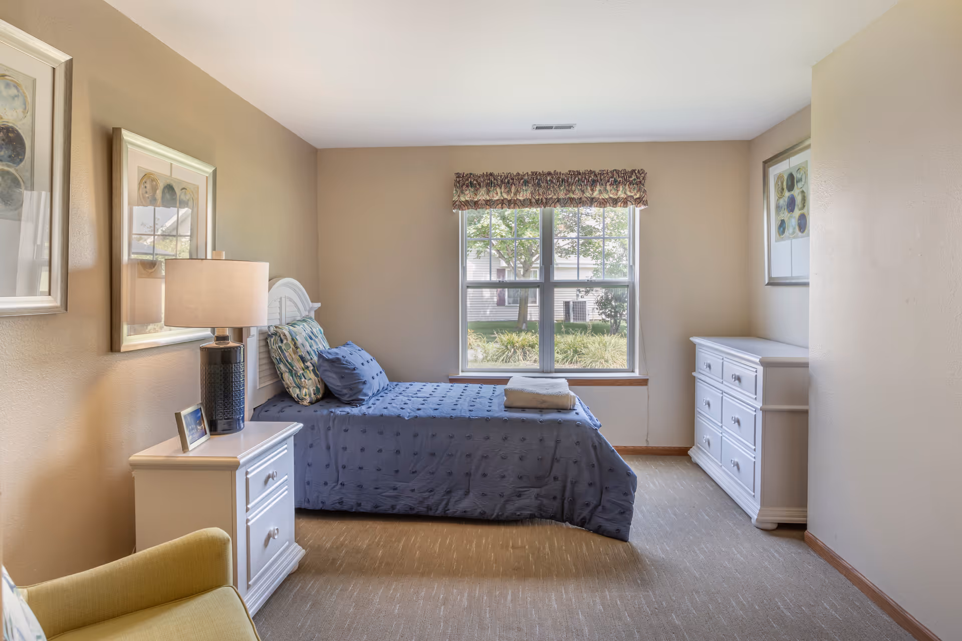 Sunlit bedroom with a single bed dressed in blue bedding, white nightstand and dresser, a lamp and a window overlooking greenery.