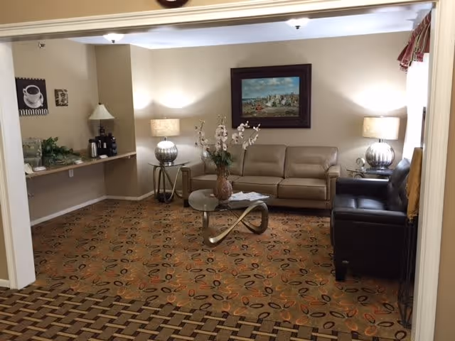 A cozy living room area with a beige leather sofa and a dark brown leather armchair. There is a modern glass coffee table with a decorative vase of flowers on it. Two side tables with matching lamps flank the beige sofa. A framed painting hangs on the wall above the sofa. To the left, there is a small counter with a lamp, some greenery, and a coffee station. The room has patterned carpet flooring and neutral-colored walls.