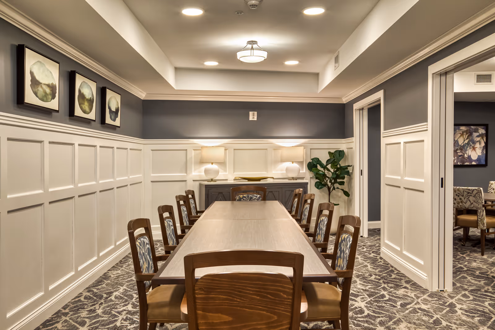 A well-lit dining room with a long wooden table surrounded by eight chairs with patterned cushions. The walls are painted gray on the upper half and white with paneling on the lower half. Three framed abstract art pieces hang on the left wall. At the far end of the room, there is a sideboard with two table lamps and a decorative plant beside it. The carpet has a floral pattern, and there is an adjacent room visible through an open doorway on the right.