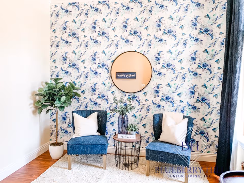 A cozy sitting area with two blue upholstered chairs each with a white pillow, a small round wireframe side table with a vase of flowers and a welcome sign, a potted plant on the left, and a round mirror on a floral patterned wall.