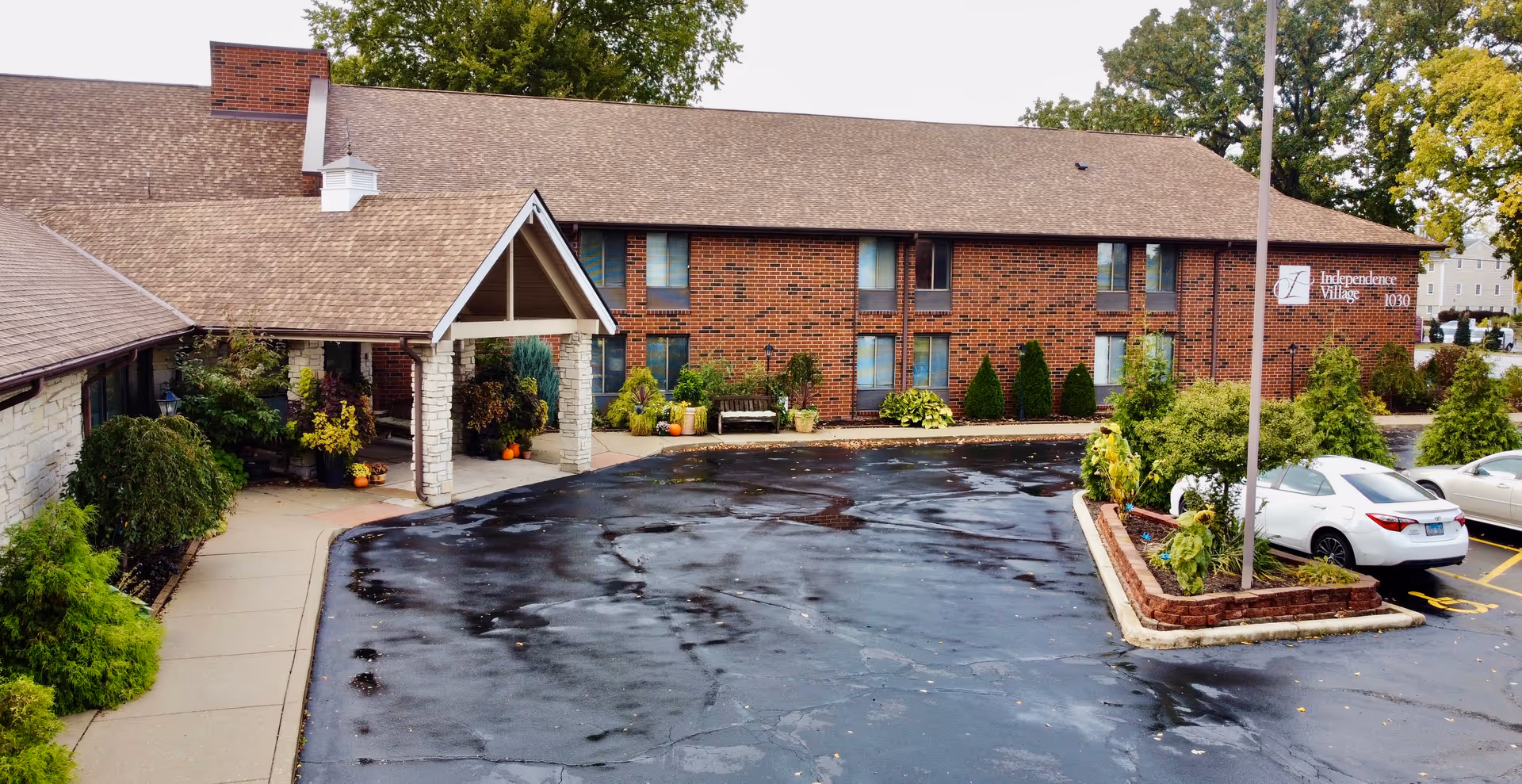 Exterior view of Independence Village senior living facility showing a brick building with multiple windows, a covered entrance with stone pillars, a parking lot with a few cars, and landscaped greenery including bushes and small trees.