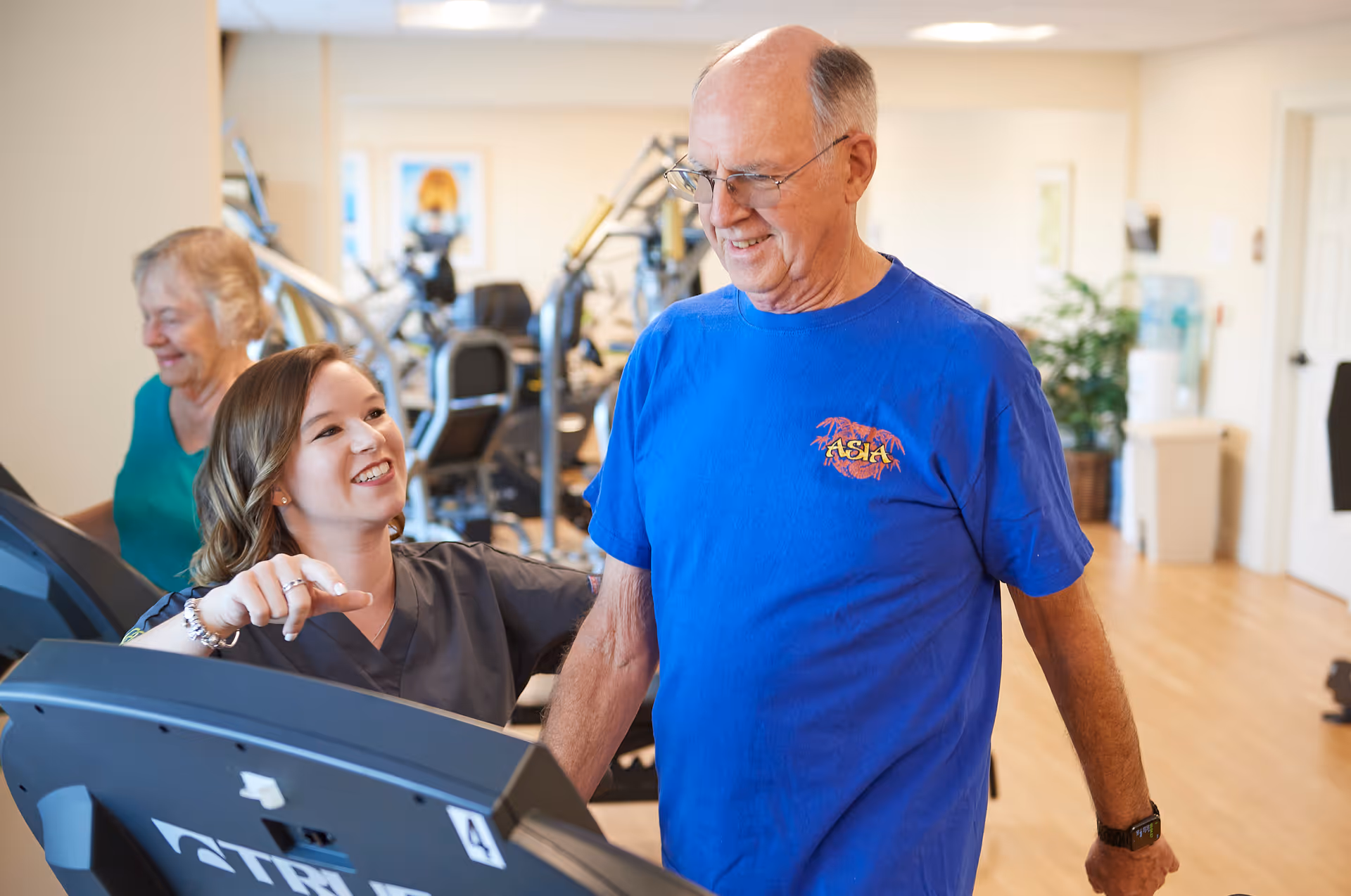 A senior man in a blue shirt is walking on a treadmill while a young female staff member in gray scrubs encourages him. In the background, an elderly woman is also using exercise equipment in a well-lit fitness room.