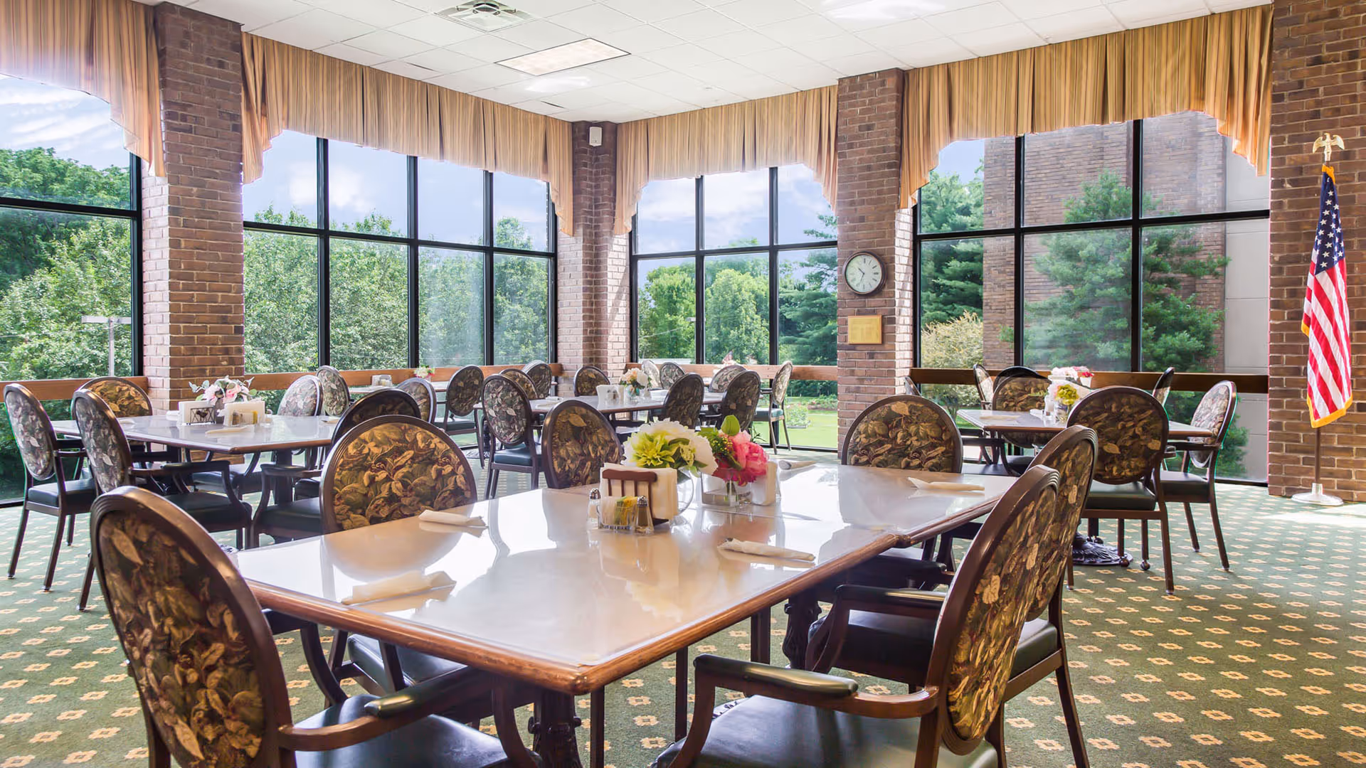 A bright dining room with large windows showing green trees outside. The room has multiple tables with floral-patterned chairs, each table set with napkins, flowers, and condiments. There is an American flag in the corner and a clock on a brick pillar.
