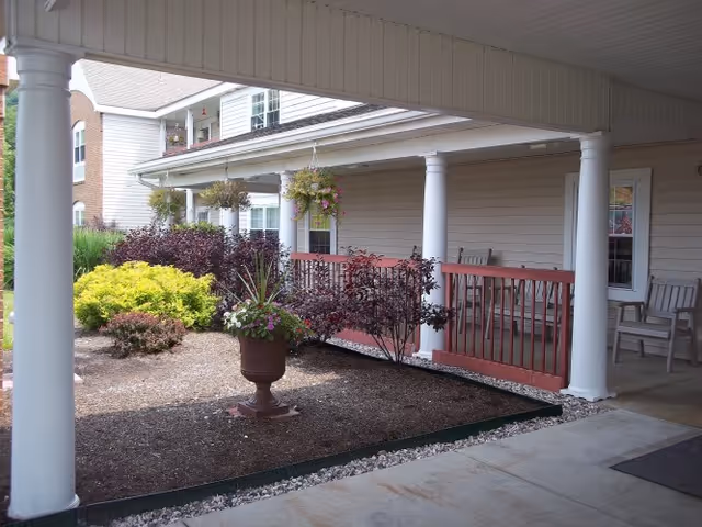 Covered porch area with white columns and red railing, overlooking a landscaped garden bed with various shrubs and a large planter with flowers. The porch has chairs and overlooks a beige building with windows and a balcony above.