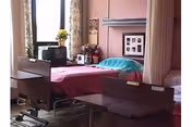 A hospital-style room with a single bed covered in a pink sheet and a blue pillow. The room has a window with curtains, a small table with flowers and framed pictures, and a brown recliner chair in the foreground.