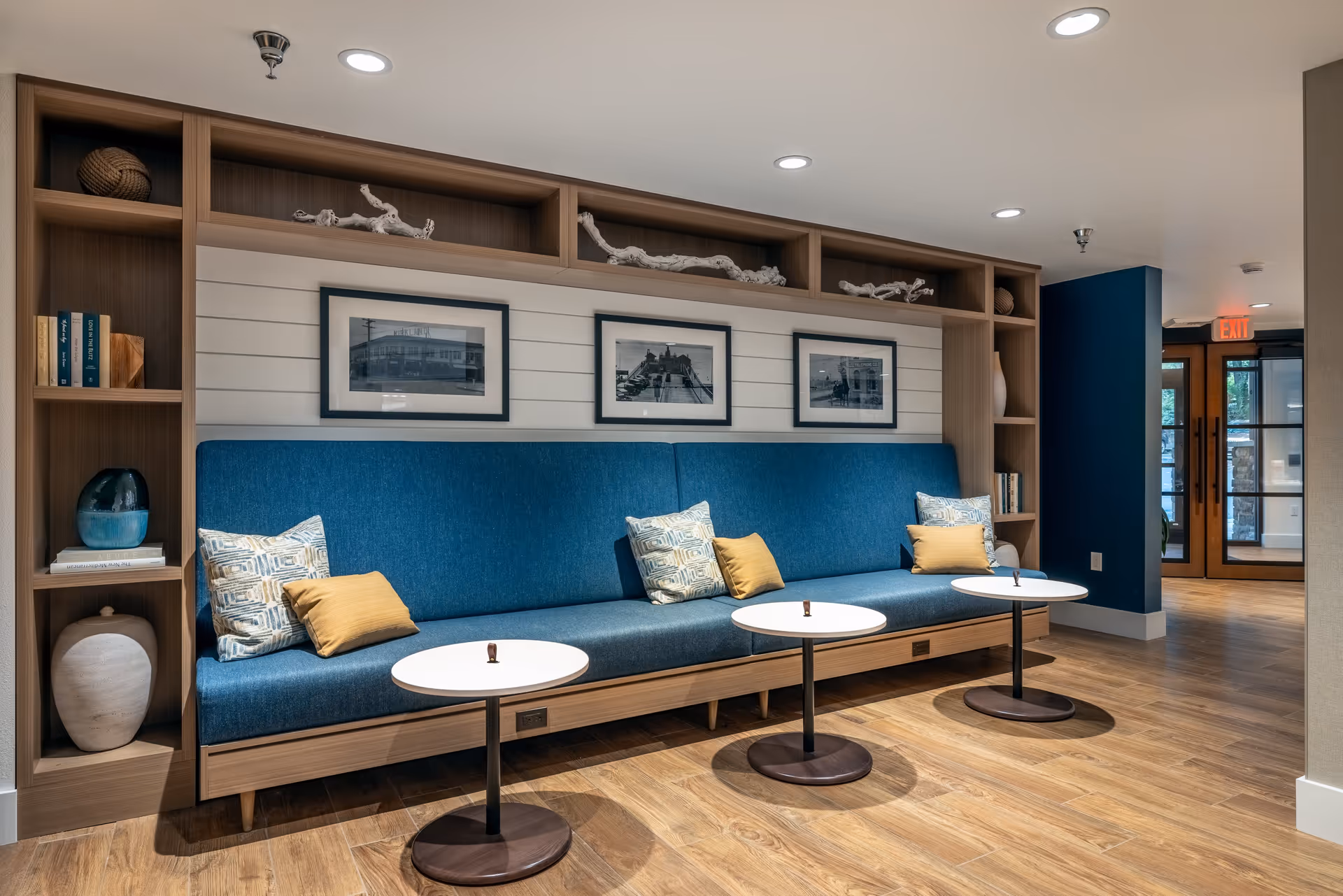 A modern lounge area with a long blue cushioned bench adorned with patterned and solid yellow pillows. Above the bench are three black-and-white framed photographs mounted on a white shiplap wall. Wooden shelves on either side hold decorative items including books, vases, and driftwood pieces. Three small round tables with dark bases are positioned in front of the bench. The floor is wood, and there is a doorway with glass doors and an exit sign in the background.