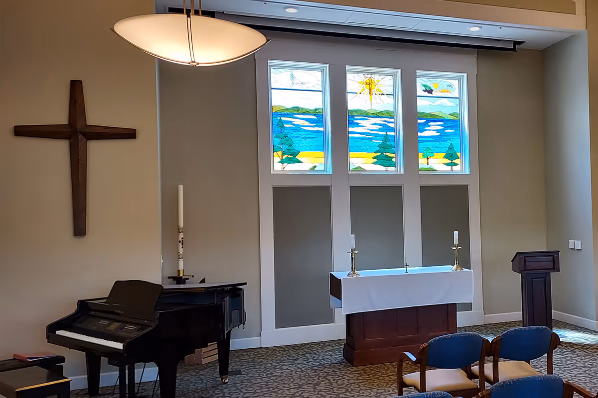 Interior of a small chapel or worship room featuring a wooden cross mounted on the wall, a black grand piano, an altar covered with a white cloth and two candlesticks, a wooden lectern, stained glass windows depicting a scenic landscape with trees, water, and a sun, and several chairs arranged in rows.