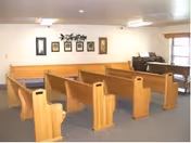 Small chapel-like interior with rows of wooden pews, framed pictures on the wall, and a piano near a window.