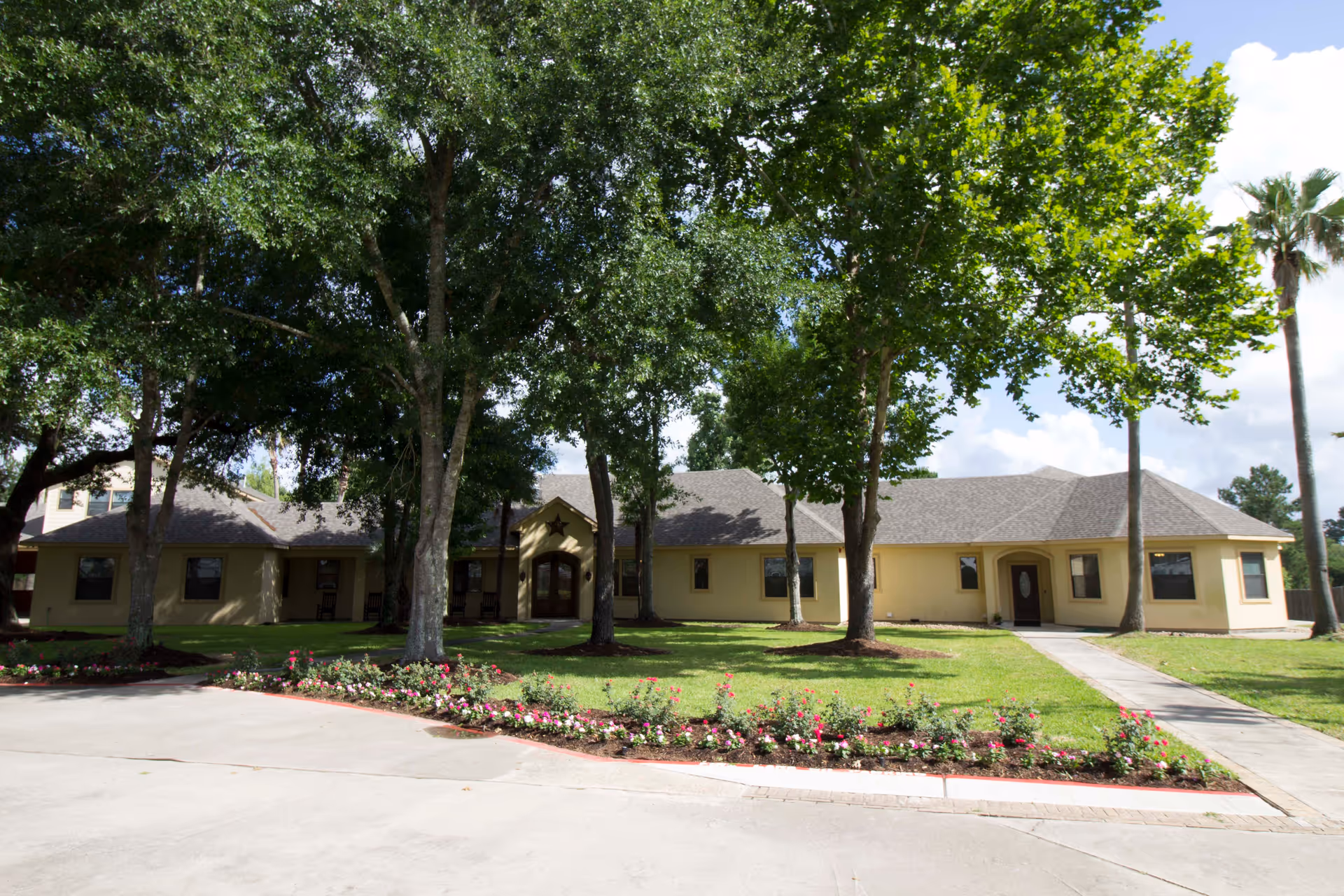 Front exterior view of a single-story senior living facility building with a beige facade, multiple windows, and two entrance doors. The building is surrounded by a well-maintained lawn, flower beds with pink and white flowers, and several tall trees providing shade. The sky is partly cloudy.