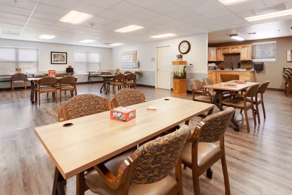 A spacious, well-lit common area with multiple tables and chairs arranged for group activities or dining. The room features wood flooring, a small kitchen area with wooden cabinets, a clock on the wall, and windows with blinds allowing natural light. Board games are placed on the tables, suggesting a recreational or social setting.