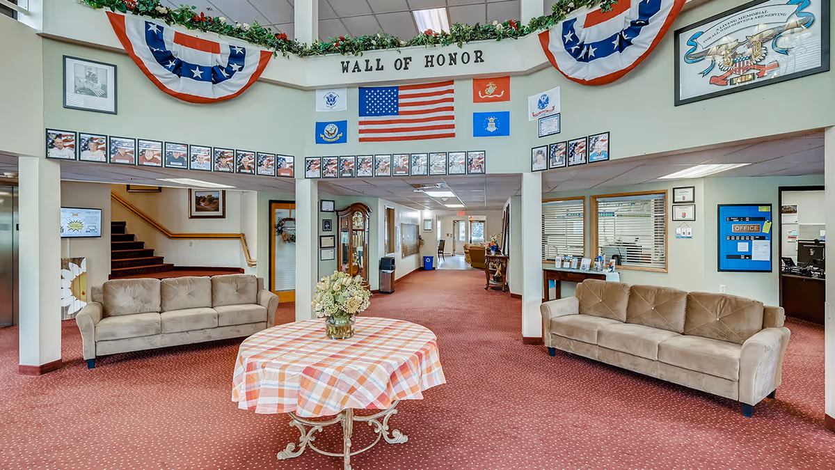 Interior view of a senior living facility lobby area with two beige sofas, a round table with a checkered tablecloth and a flower arrangement in the center. Above the entrance is a 'Wall of Honor' display featuring an American flag, military branch flags, and framed photos of veterans. The space has red carpet flooring, light green walls, and patriotic bunting decorations near the ceiling.
