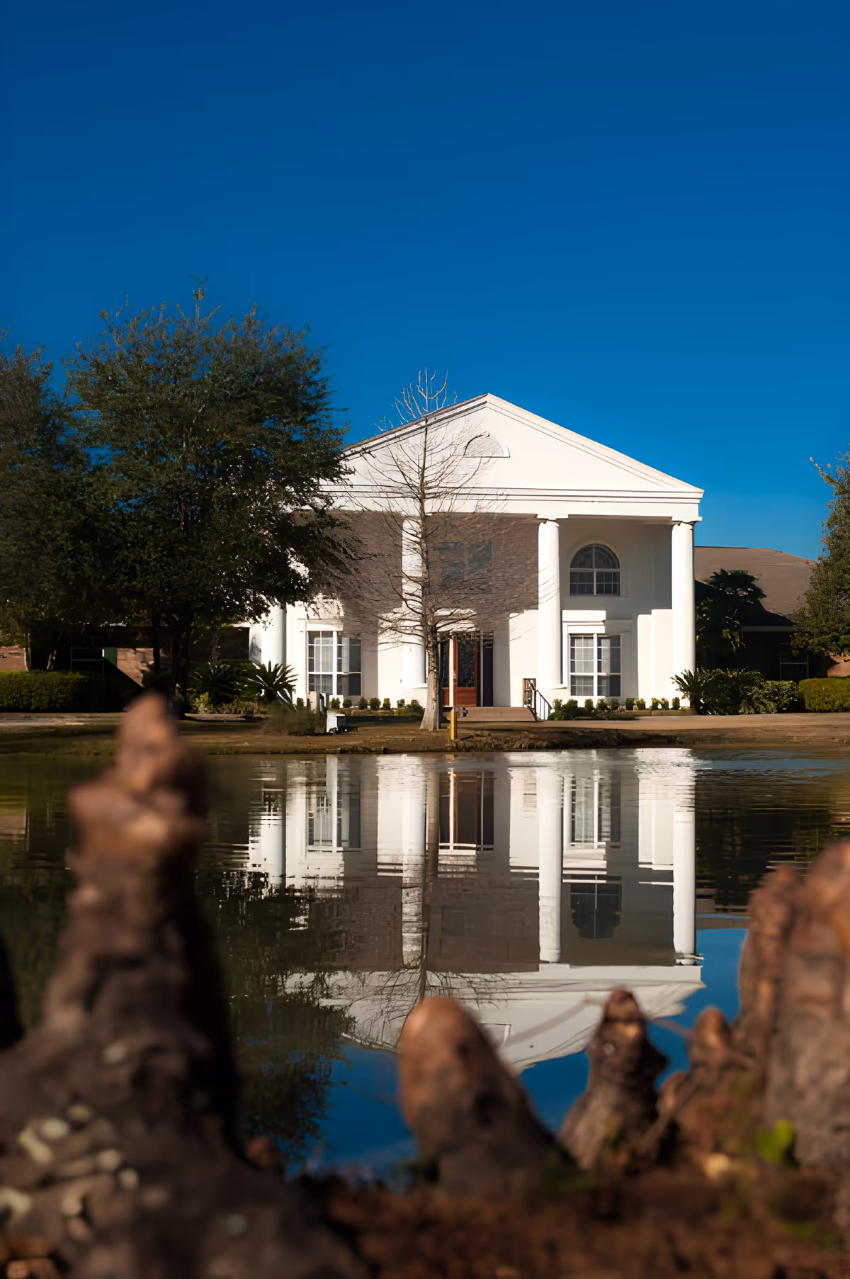 White neoclassical building with columns reflected in a pond under a clear blue sky.