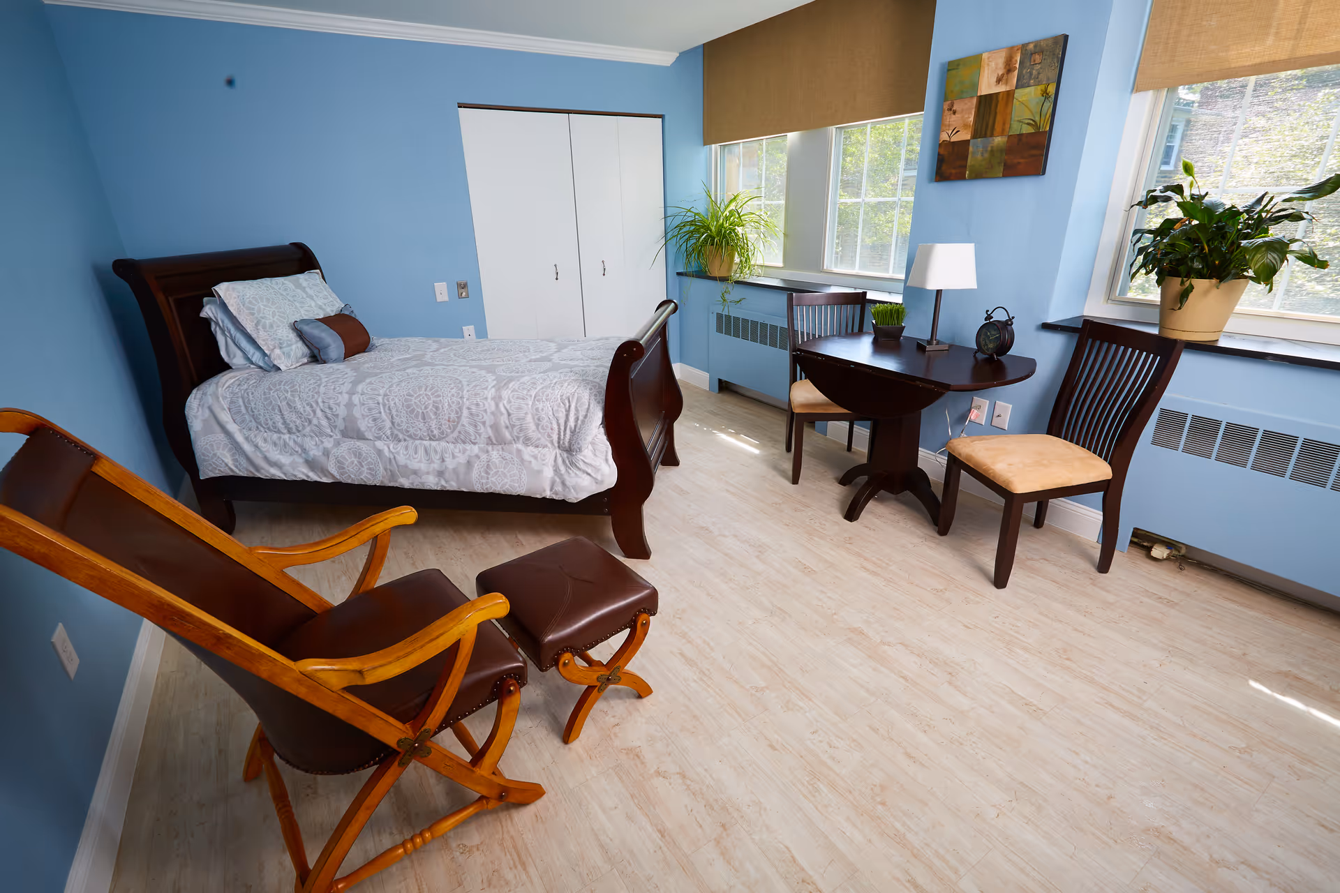 A bright bedroom with light blue walls and light wood flooring featuring a dark wooden bed with white patterned bedding, a wooden rocking chair with a matching footrest, a small round dark wooden table with two chairs, a table lamp, an alarm clock, and two potted plants on the windowsill.