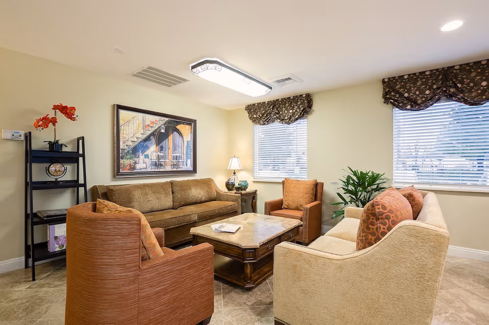 A cozy living room with a brown sofa, two orange armchairs, and a beige sofa arranged around a wooden coffee table. The room has two windows with floral valances, a black shelving unit with decorative items including a clock and red flowers, a table lamp on a side table, and a framed painting of an outdoor staircase on the wall.