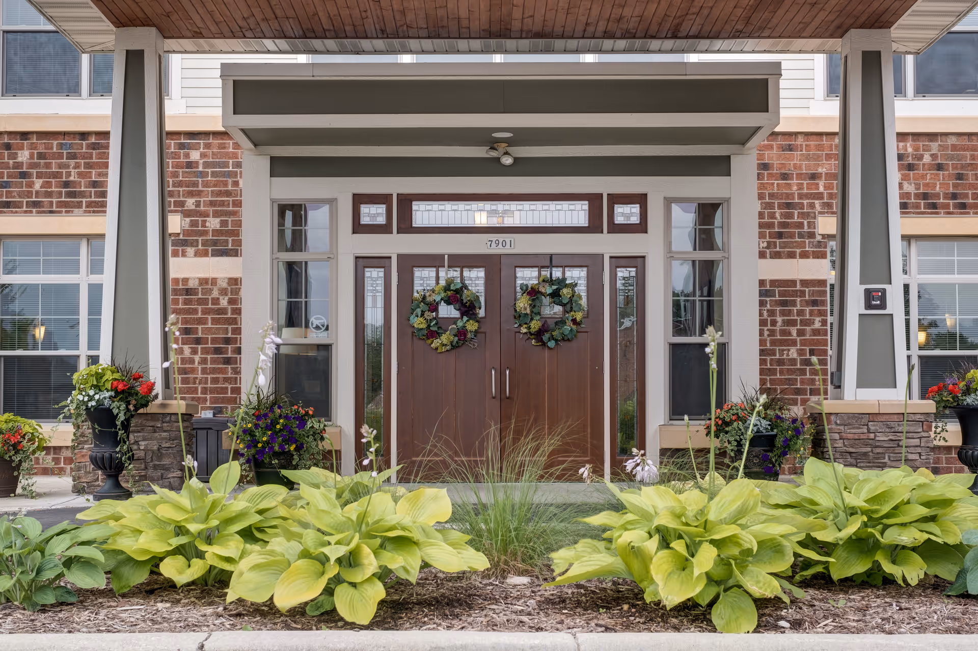 Front entrance of a brick building with double wooden doors decorated with floral wreaths. The entrance is framed by large windows and columns, with green plants and colorful flowers in planters on either side and a garden bed with leafy plants in front.
