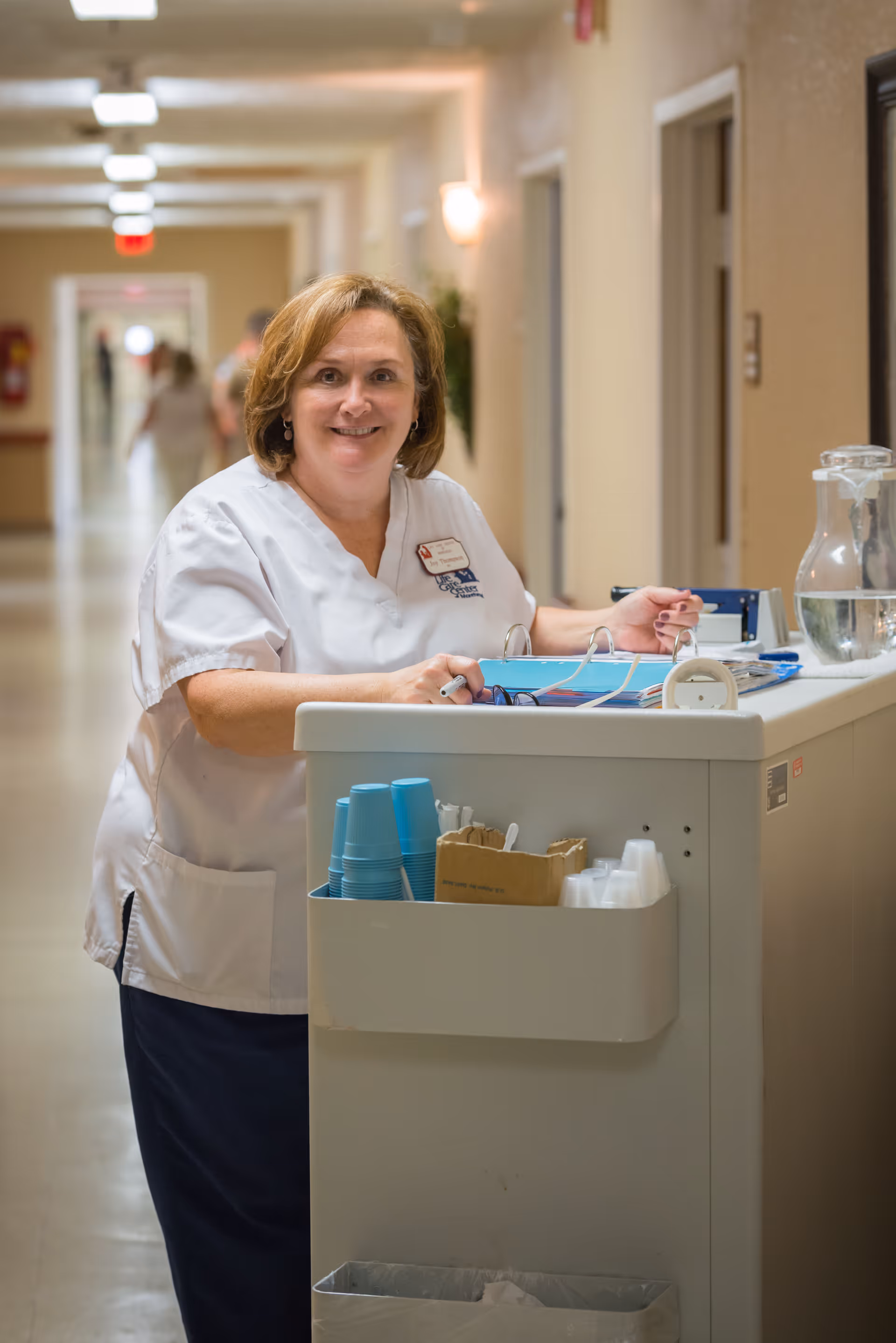 A smiling female healthcare worker in a white uniform stands behind a medical cart in a well-lit hallway of a senior living facility. The cart holds blue disposable cups, a box with utensils, a water pitcher, and some paperwork. The hallway has beige walls and several doors along it.