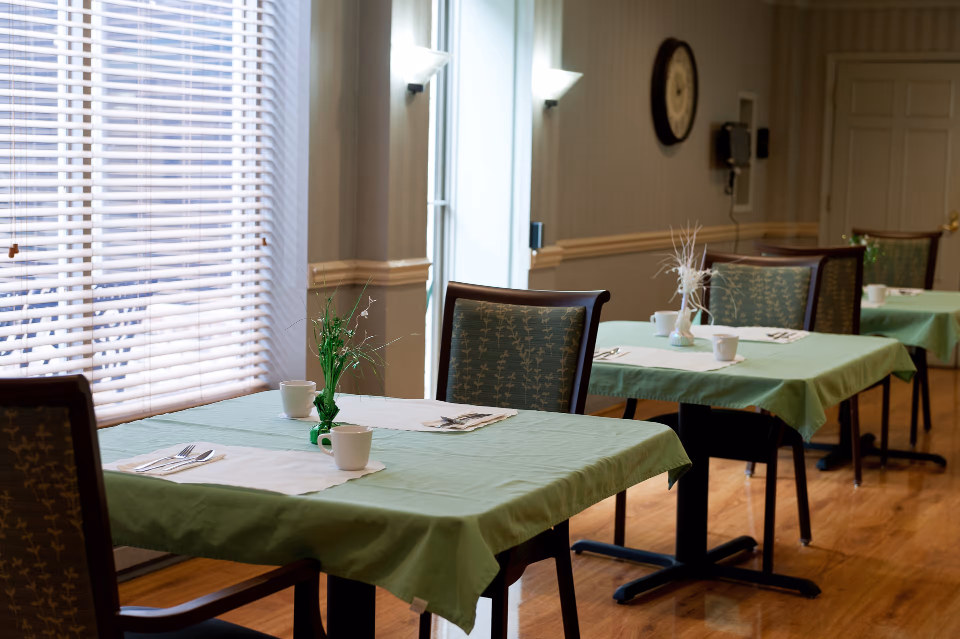 A dining area with tables covered in green tablecloths, each set with white placemats, cups, utensils, and small decorative plants. The room has wooden floors, a window with blinds, and a wall clock.
