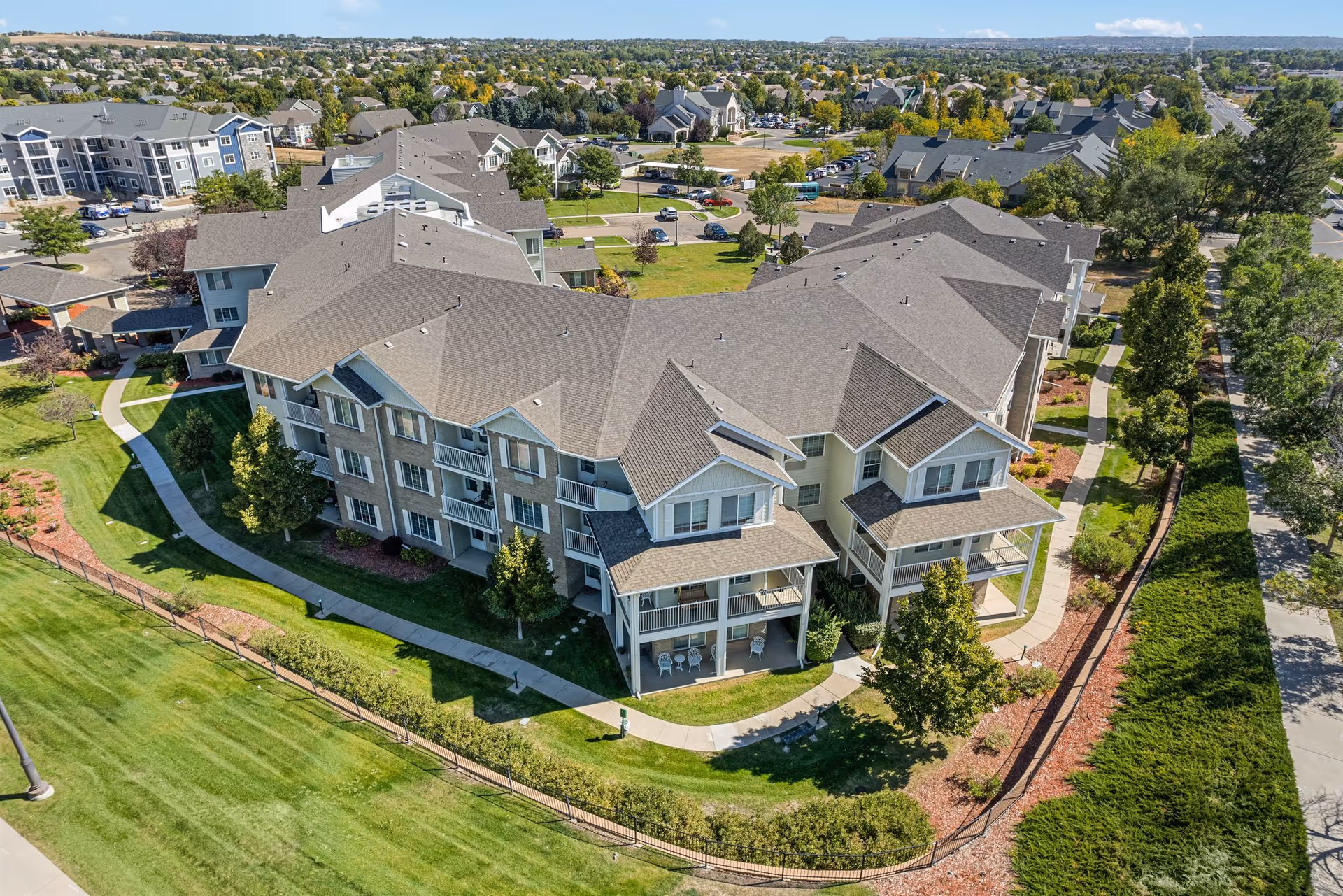 Aerial view of a large residential building complex with multiple units, surrounded by green lawns, trees, and paved walkways. The building has multiple balconies and a gray shingled roof. The surrounding neighborhood includes other residential buildings and streets.