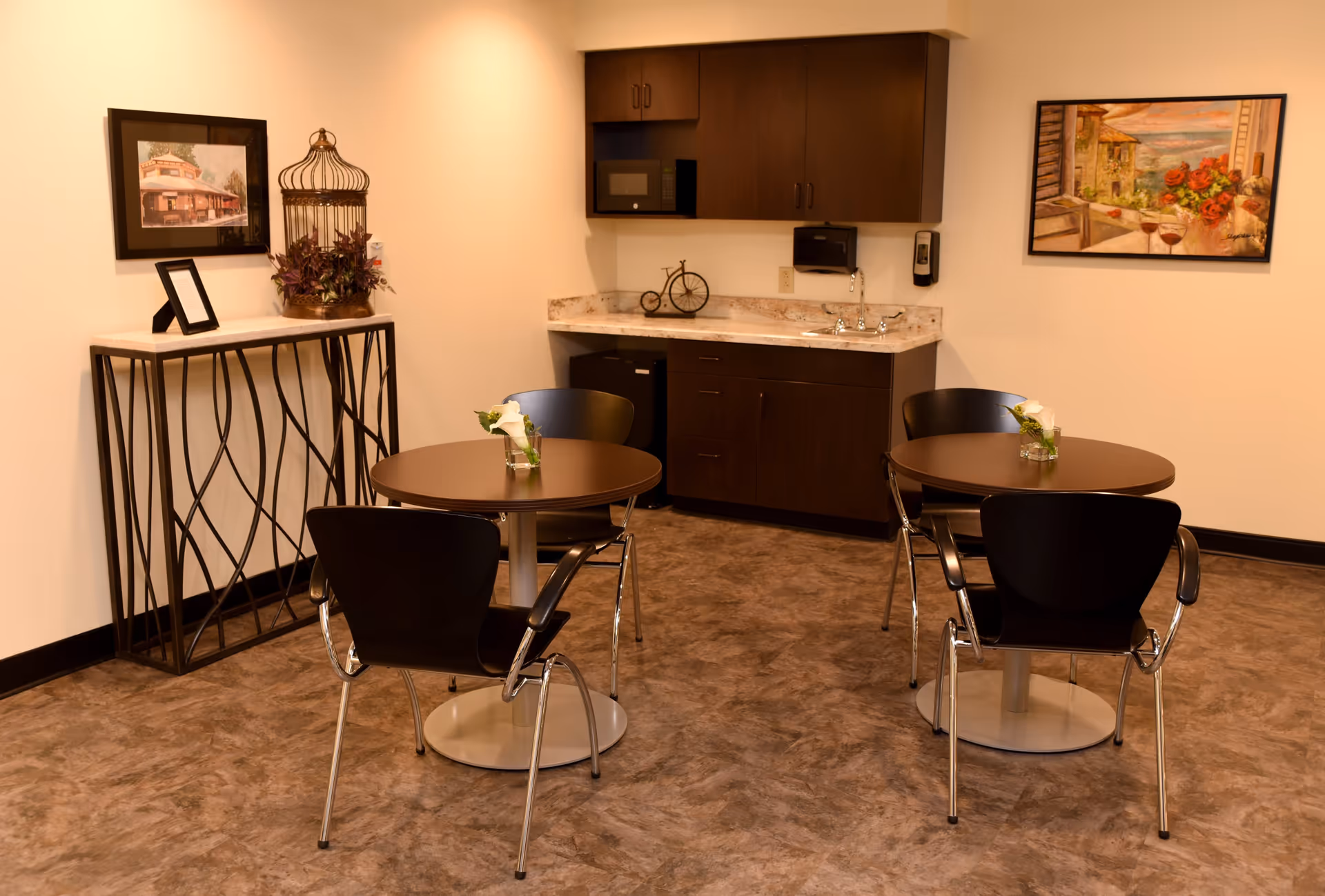 A small dining area with two round tables, each surrounded by three black chairs. Each table has a small vase with white flowers. In the background, there is a kitchenette with dark wood cabinets, a microwave, a sink, and a small refrigerator. The walls are decorated with two framed paintings, and there is a decorative console table with a birdcage and plants on the left side.