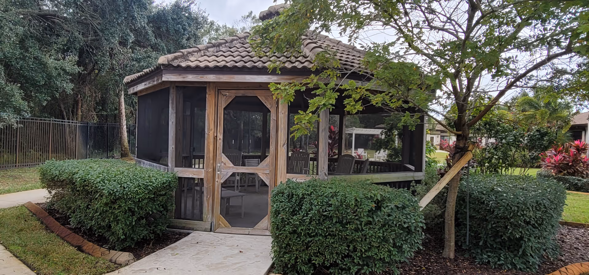 A wooden gazebo with a tiled roof and screened windows, surrounded by green bushes and trees, located in an outdoor garden area of a senior living facility.