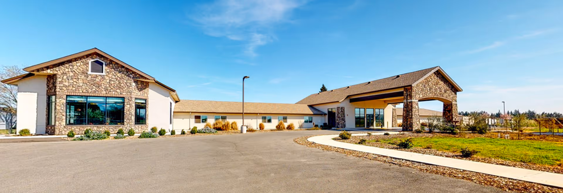 Exterior view of a single-story building with stone and beige siding, featuring a covered entrance with stone pillars, a large window, and a paved driveway surrounded by landscaped greenery under a clear blue sky.