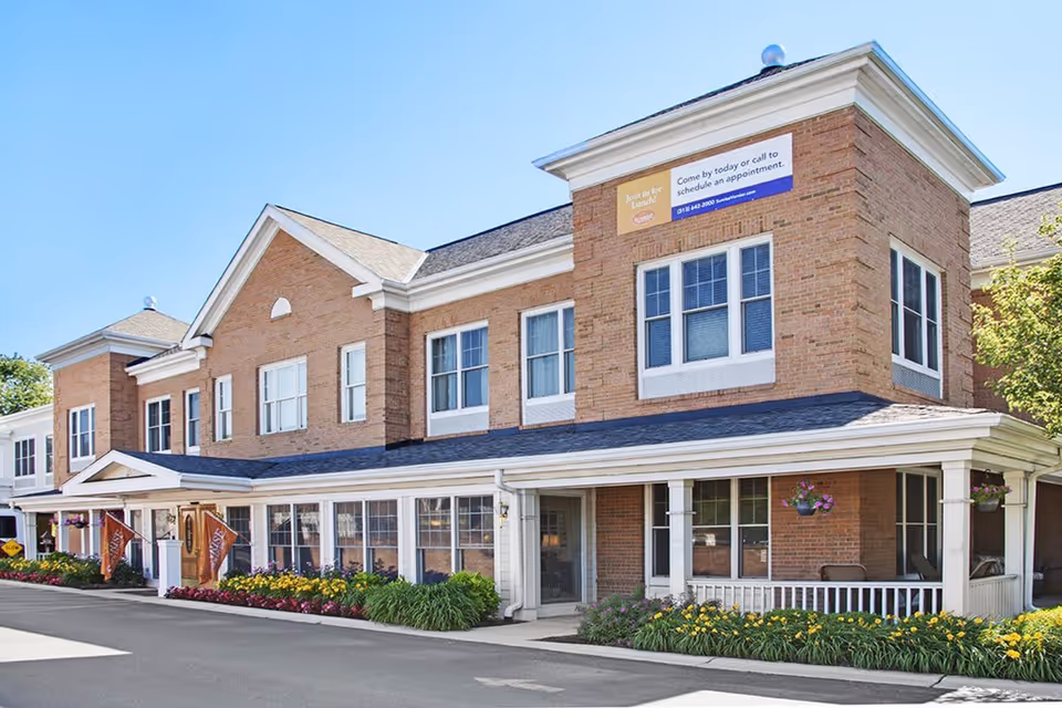 Exterior view of a two-story brick senior living facility building with white trim, multiple windows, a covered porch with hanging flower baskets, and landscaped flower beds along the front. The sky is clear and blue.