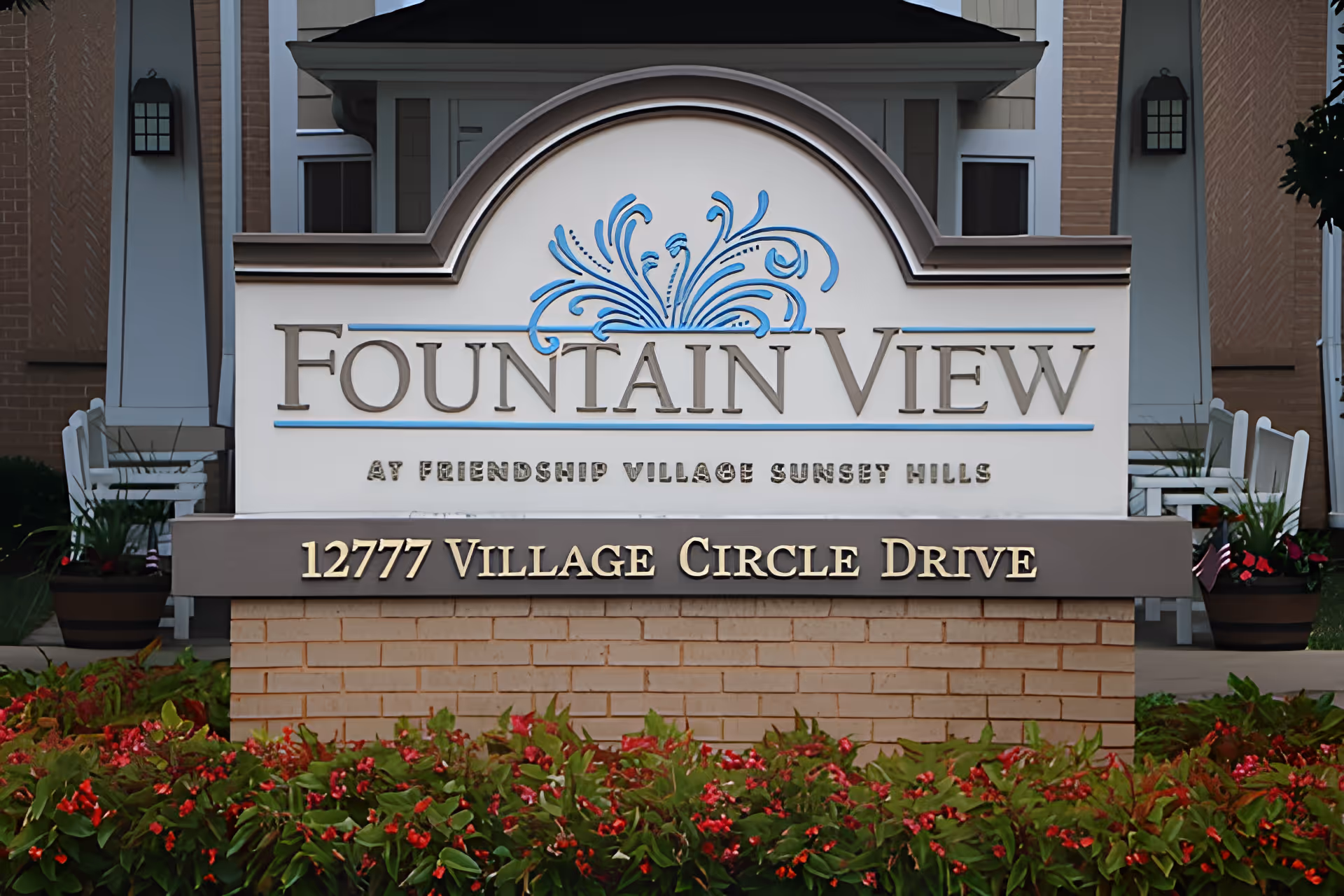Entrance sign for Fountain View at Friendship Village Sunset Hills, displaying the address 12777 Village Circle Drive, surrounded by flowers and part of the building exterior in the background.