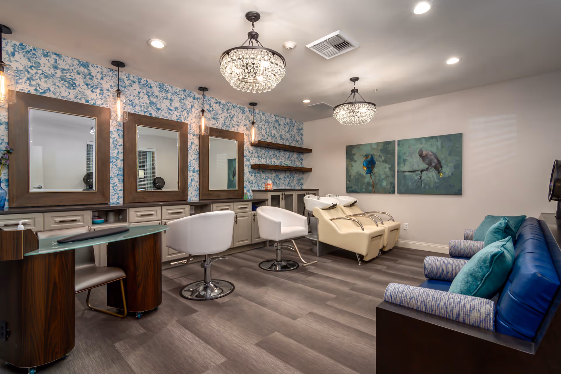 Interior of a senior living facility salon with three large mirrors mounted on a floral wallpapered wall, two white salon chairs, two cream-colored shampoo stations, a dark wood manicure table with a chair, and a blue cushioned bench with teal and patterned pillows. The room is lit by two crystal chandeliers and recessed ceiling lights, with two paintings of birds on the far wall.