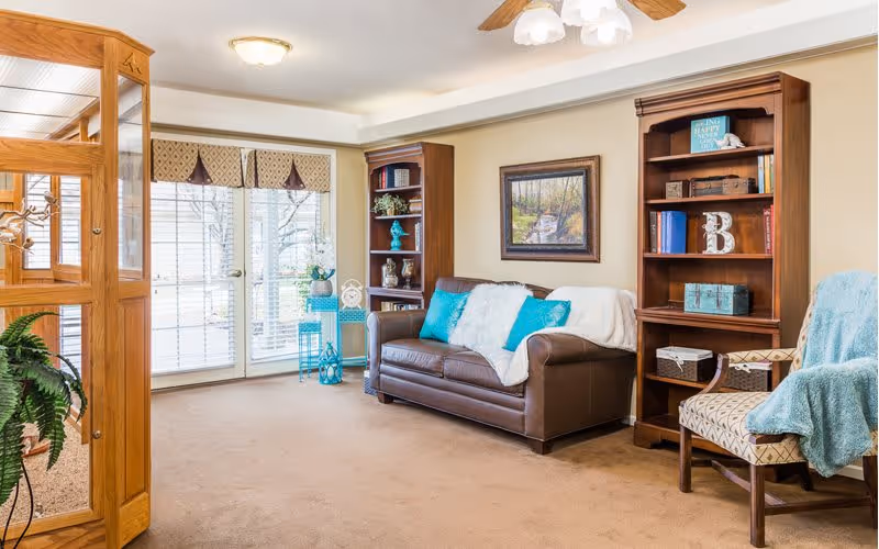 A cozy living room with a brown leather sofa adorned with white and turquoise pillows and a white throw blanket. There are two wooden bookshelves filled with books and decorative items on either side of the sofa. A patterned armchair with a turquoise throw blanket is positioned to the right. Large windows with blinds and a valance allow natural light to fill the room. A wooden display cabinet with glass doors and a green potted plant are visible on the left side.