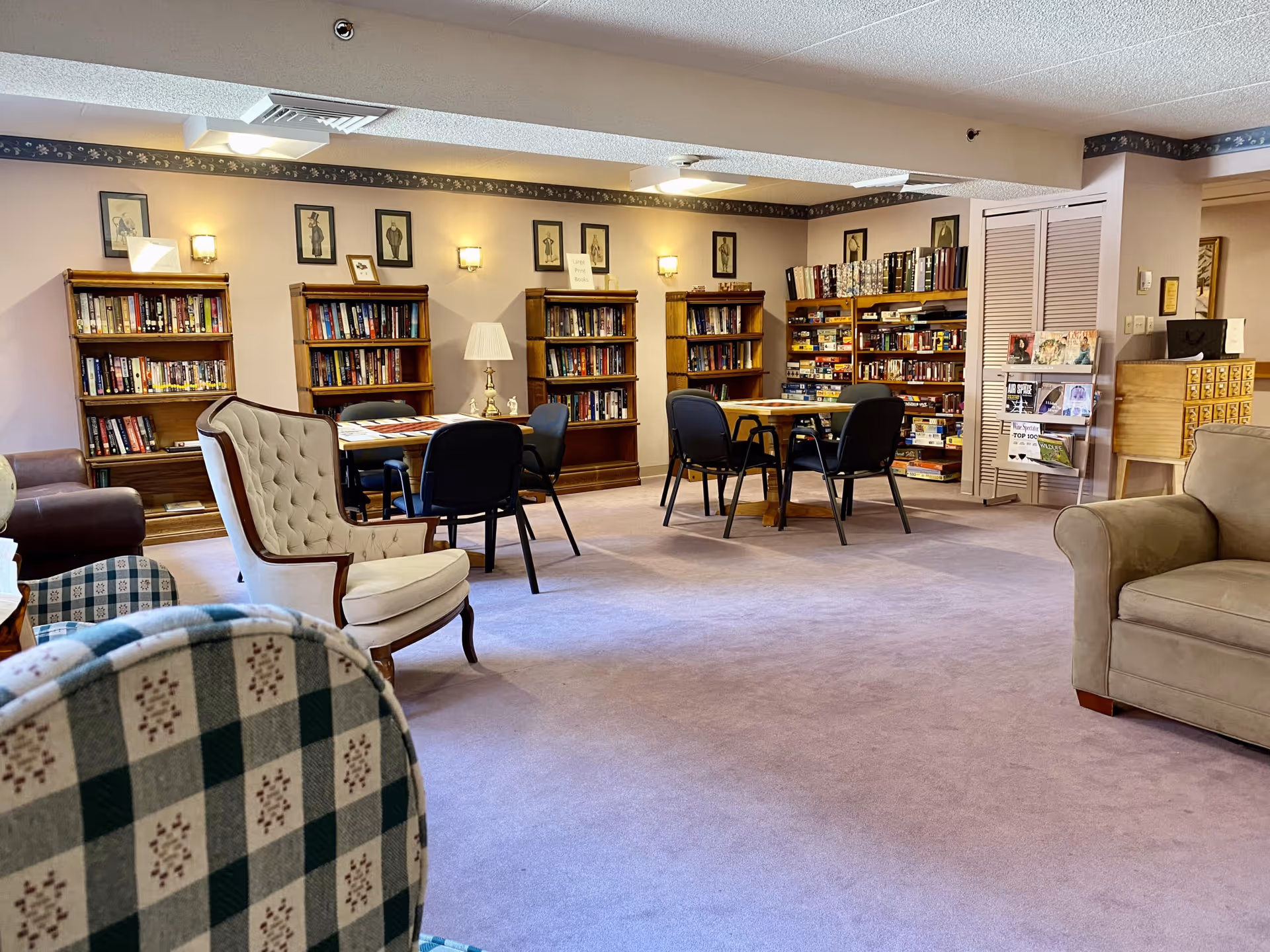 A cozy library or common room in a retirement community with bookshelves filled with books and board games, several tables with chairs, and comfortable armchairs. The room has soft lighting and framed artwork on the walls.