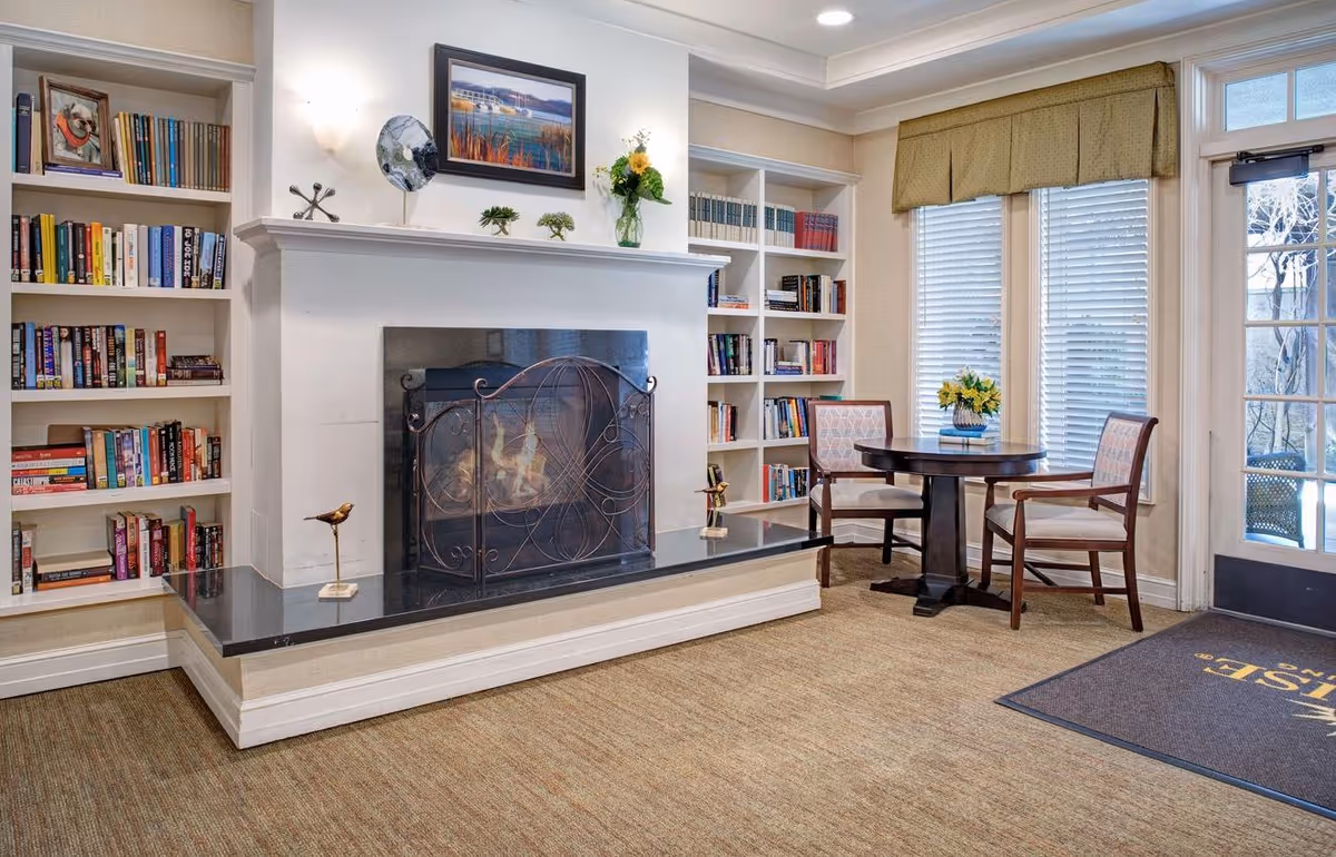 Cozy common room with a lit fireplace flanked by built-in bookshelves, a small round table with two chairs by windows, and a door mat near the entrance.