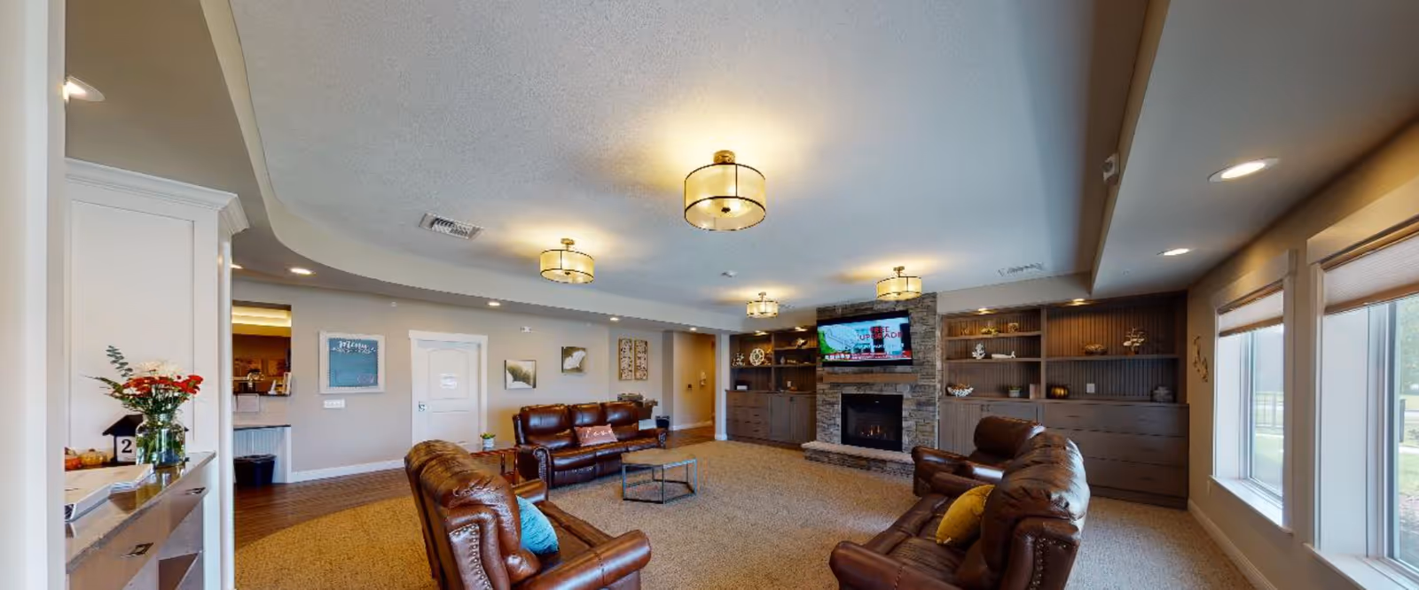 Spacious senior living lounge with leather sofas arranged around a fireplace and wall-mounted TV, built-in shelving, and large windows.