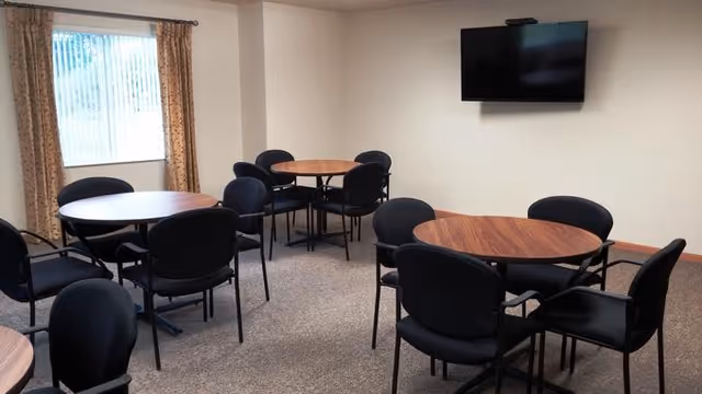 A small common area with several round wooden tables surrounded by black chairs. There is a window with patterned curtains on the left wall and a flat-screen TV mounted on the right wall. The room has beige walls and carpeted flooring.