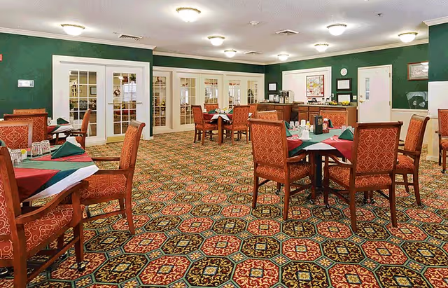 A dining room with multiple tables covered with green and red tablecloths, each set with glasses and napkins. The room has patterned carpet flooring, green walls, and white-framed glass doors along one side. Ceiling lights illuminate the space, and there is a counter area with coffee machines and other items in the background.