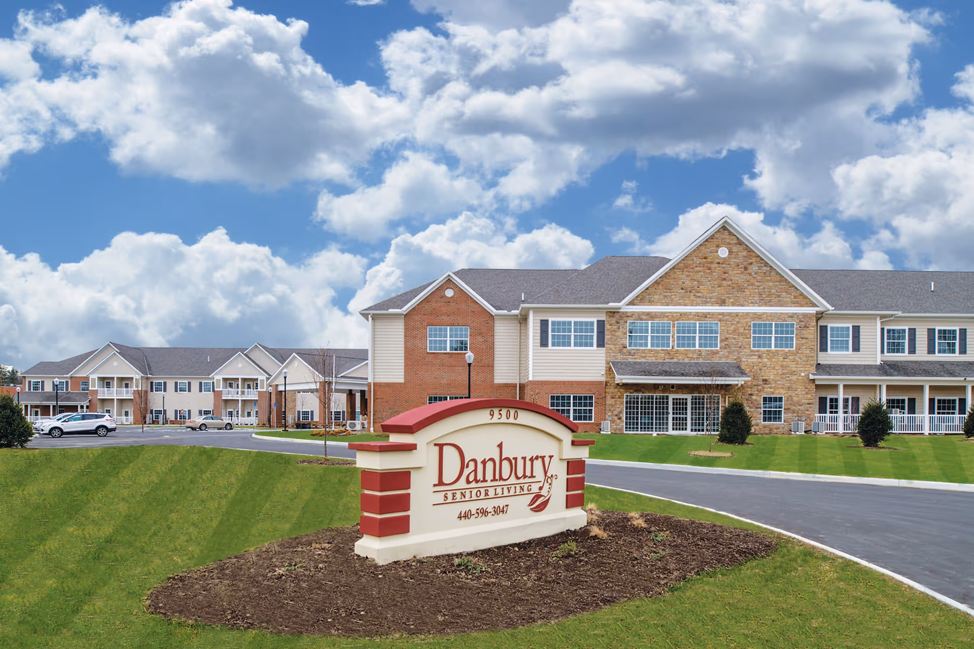 Exterior view of Danbury Broadview Heights senior living facility with a large sign in the foreground displaying the name and phone number. The building is two stories with a mix of brick and siding, surrounded by a well-maintained lawn and a paved driveway under a partly cloudy sky.