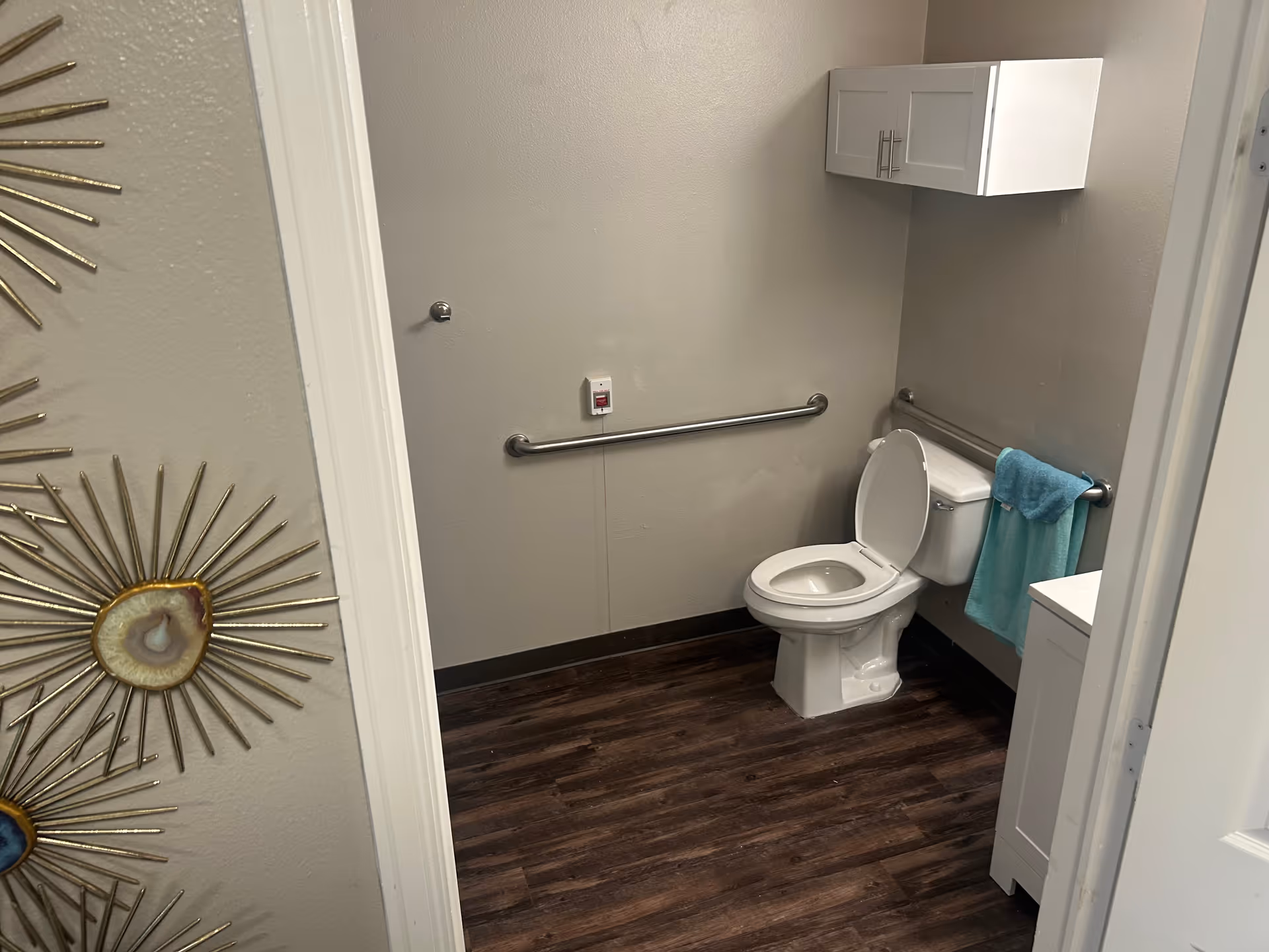 View into a small bathroom with a white toilet that has the lid open, a white cabinet mounted on the wall above the toilet, a towel bar with a blue towel, a grab bar on the wall, and a wooden floor. Part of a decorative wall piece with gold and stone elements is visible on the left side of the image.