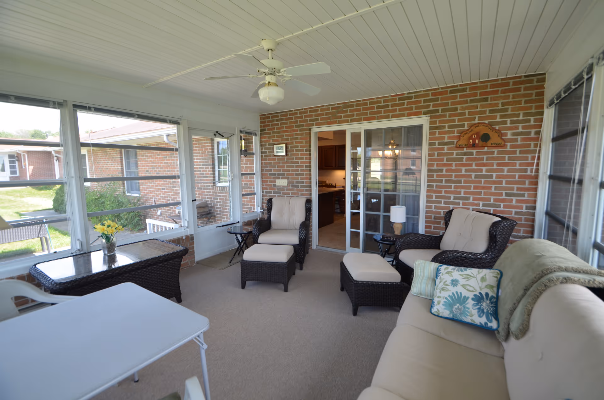 A bright enclosed porch area with large windows and a ceiling fan. The space features a beige cushioned sofa with decorative pillows and a throw blanket, two cushioned armchairs with matching ottomans, a small side table with a lamp, and a wicker table with a vase of yellow flowers. The walls are brick, and sliding glass doors lead to an interior room with a chandelier visible inside.