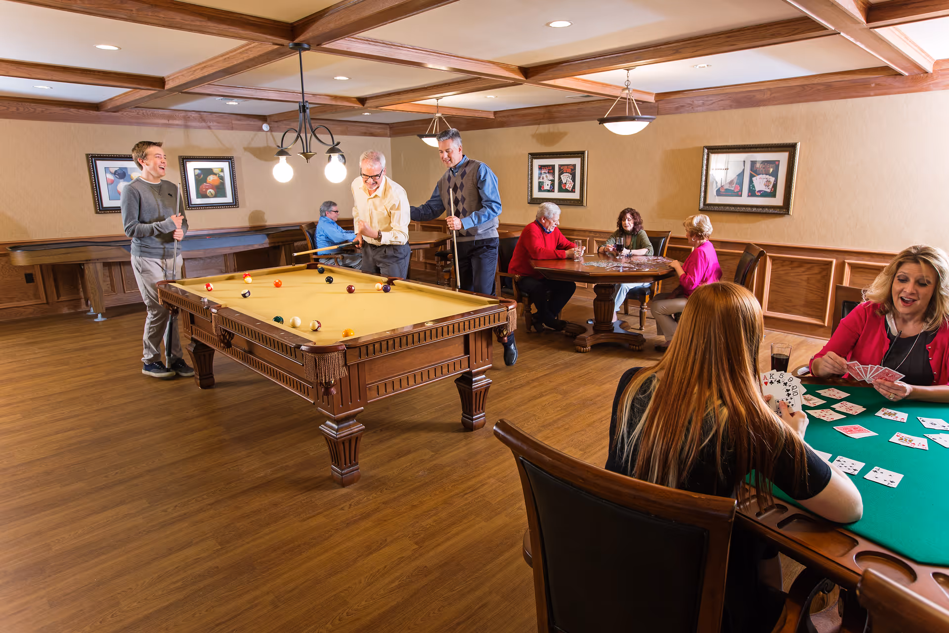 A group of seniors enjoying recreational activities in a community room. Three men are playing pool on a billiards table, while four women are seated at two tables playing card games. The room has wooden flooring, beige walls with framed artwork, and a coffered ceiling with hanging lights.