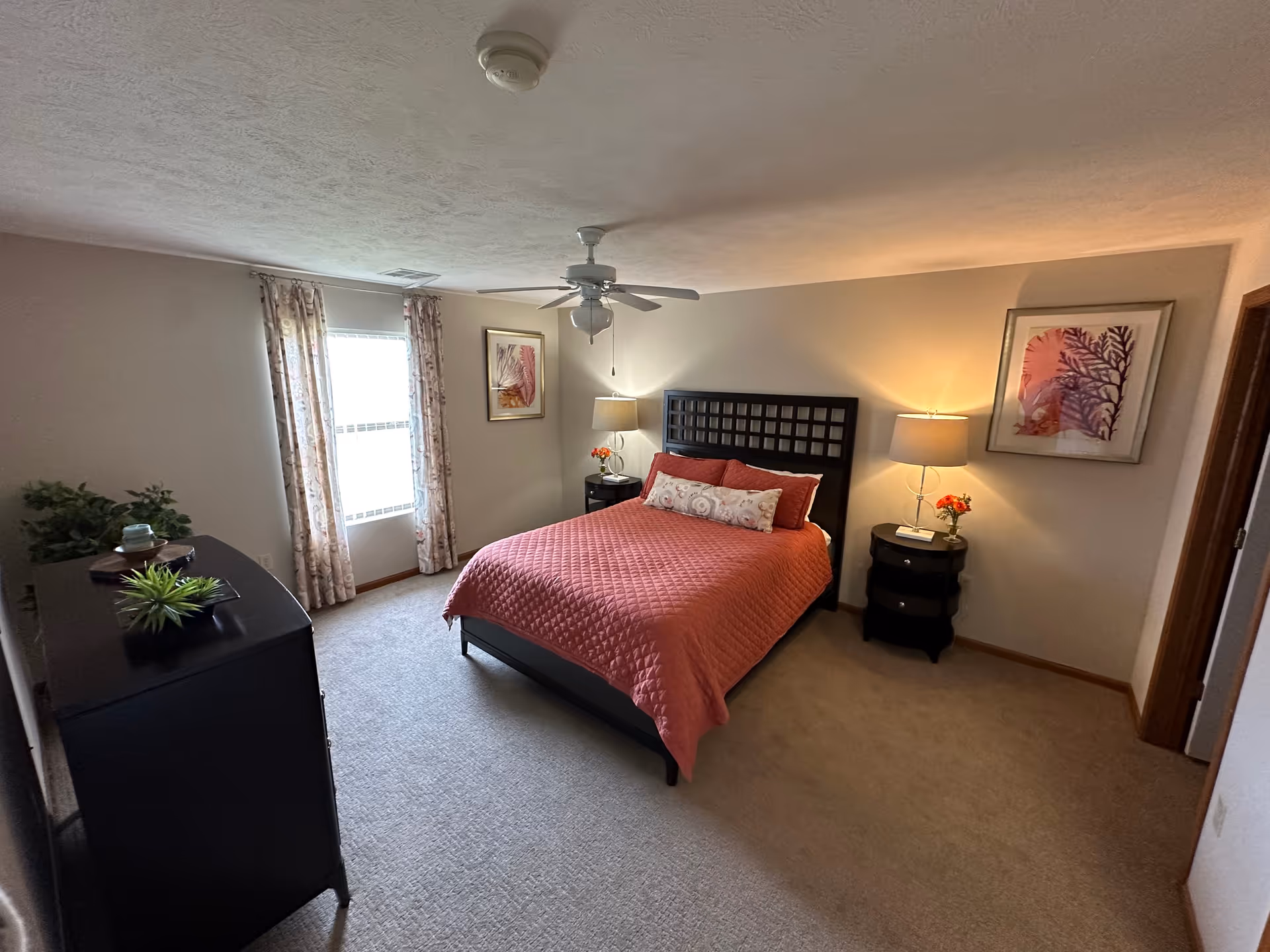 A bedroom with a queen-sized bed covered in a coral quilt and decorative pillows. The room has beige carpet, two nightstands with lamps and flowers, a ceiling fan, a window with floral curtains, framed artwork on the walls, and a black dresser with plants and decorative items.