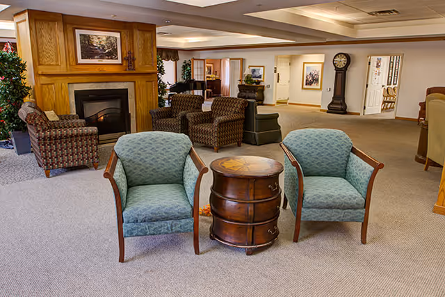 A cozy senior living facility lounge with two upholstered chairs and a small round wooden table in front of a fireplace and additional seating areas.