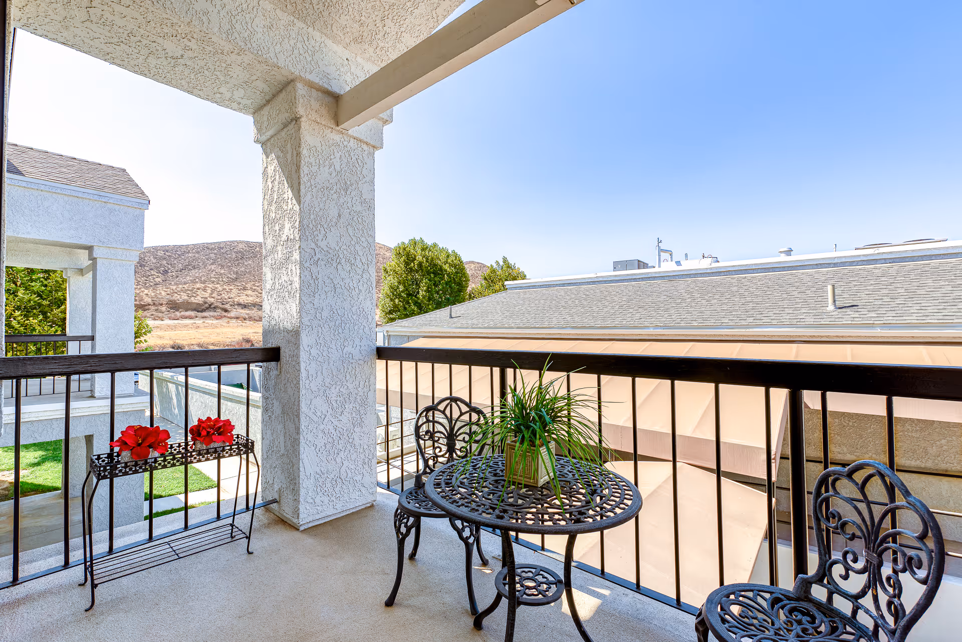 Covered balcony with a small wrought-iron table and chairs, a potted plant and flower stand overlooking neighboring rooftops and distant hills.