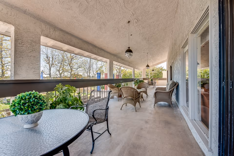 Covered outdoor patio area with multiple seating arrangements including wicker chairs and a small round table with a potted plant. The patio overlooks trees and a garden area, with hanging pendant lights overhead.