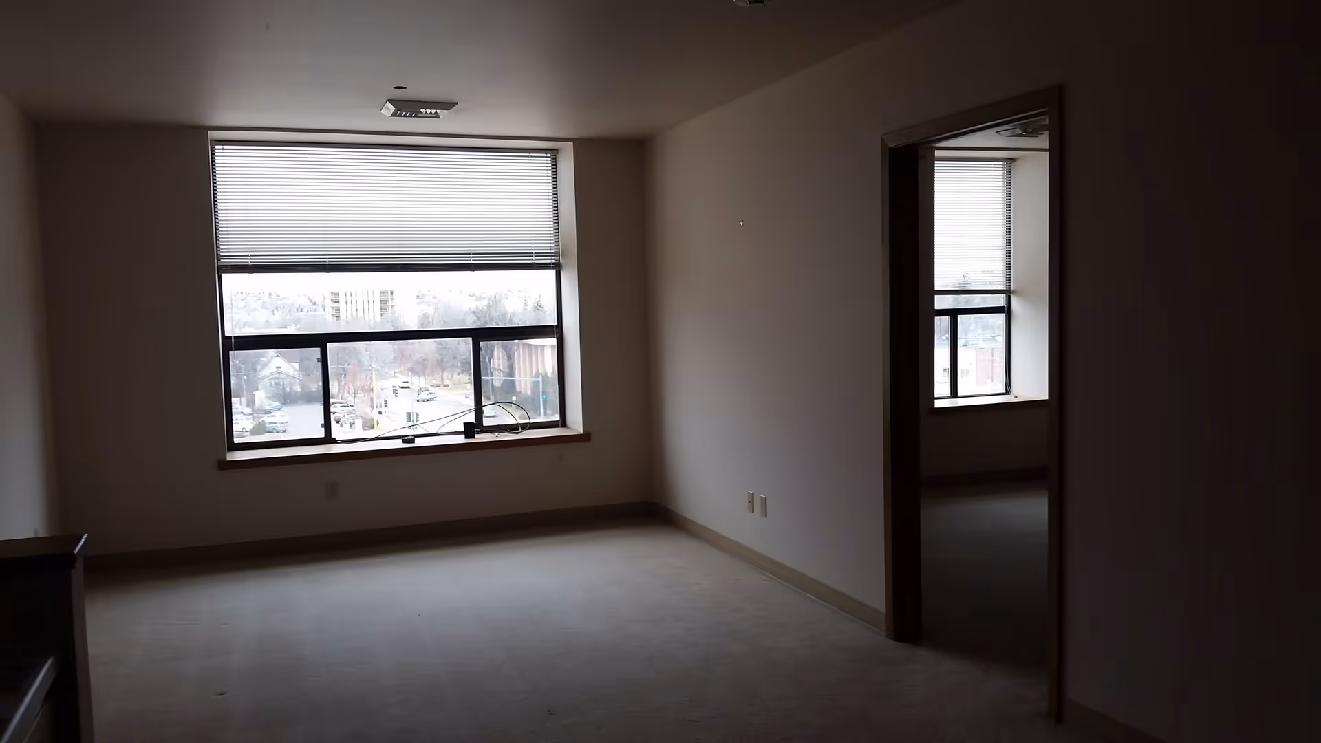 Empty room with beige carpet and walls, a large window with blinds partially open showing an outside view of buildings and trees, and an open doorway leading to another room with a similar window.